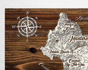 May include: A wooden map of Monterey, California, with a compass rose in the upper left corner. The map shows the city's streets and landmarks, including Carmel-by-the-Sea, Pebble Beach, and Del Ray Oaks. The text "Papier Blanc" is printed in the lower left corner.