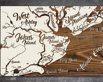 Könnte beinhalten: Eine weiße Holzkarte der Gegend von Charleston, South Carolina, mit den Namen verschiedener Inseln und Orte, darunter Johns Island, James Island, West Ashley, Mount Pleasant, Isle of Palms, Folly Beach, Kiawah Island und Sullivan's Island. Die Karte befindet sich auf einem braunen Holzgrund mit einer Kompassrose unten rechts.