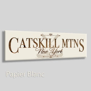 May include: A white rectangular sign with brown text that reads "CATSKILL MTNS New York". The sign features a stylized image of two crossed paddles with a canoe in the center.