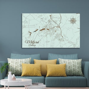 May include: A light blue wooden map of Milford, Delaware with a compass rose in the top right corner. The map is mounted on a wall above a teal couch with yellow and green patterned throw pillows.