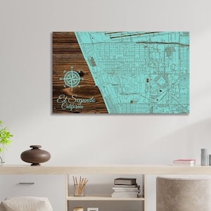 May include: A wooden wall art piece featuring a map of El Segundo, California. The map is outlined in blue on a light blue background. A compass rose and the words "El Segundo, California" are on the left side of the map.