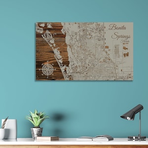 May include: A wooden map of Bonita Springs, Florida, with a compass rose in the lower left corner. The map is on a light brown wood background with a white outline of the city.