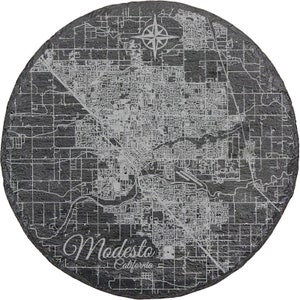 May include: A black slate coaster with a white engraved map of Modesto, California. The map includes street names and a compass rose. The text "Modesto, California" is also engraved on the coaster.