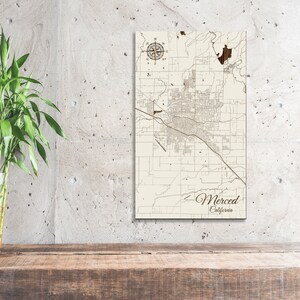 May include: A wooden map of Merced, California, with a compass rose in the top left corner. The map is mounted on a white background and has a brown wooden frame.