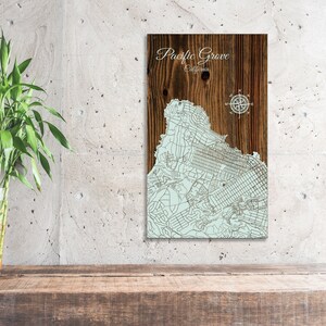 May include: A wooden map of Pacific Grove, California, with a compass rose in the top right corner. The map is on a brown wooden background.