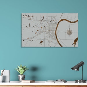 May include: A wooden map of Atchison, Kansas, with detailed streets and landmarks. The map is in shades of gray and brown, with the city name and state at the top. A compass rose is in the upper right corner.