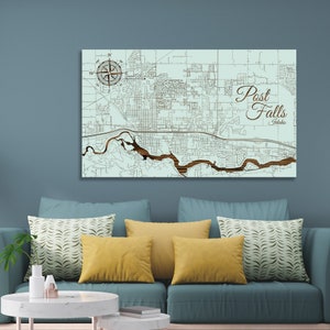 May include: A wooden map of Post Falls, Idaho, with a compass rose in the top left corner. The map is on a light blue background and the city streets are carved into the wood. The city name is written in a decorative font in the top right corner.