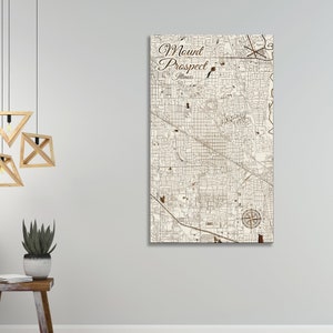 May include: A beige wood map of Mount Prospect, Illinois, with brown lines showing the streets and a compass rose in the lower right corner. The words "Mount Prospect, Illinois" are written in brown cursive at the top of the map.