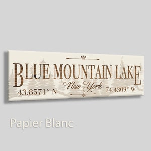 May include: A white canvas sign with brown text that reads "Blue Mountain Lake, New York 43.8571° N 74.4309° W". The sign has a rustic design with a silhouette of pine trees in the background.