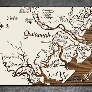 May include: A white wooden map of the Savannah, Georgia area with the names of the islands and waterways labeled in brown. The map is surrounded by a brown wooden frame.