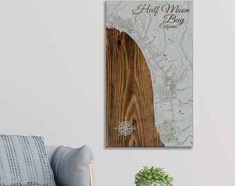 May include: A wooden map of Half Moon Bay, California, with a compass rose in the lower left corner. The map is engraved with a light gray outline of the city and surrounding area. The wood has a dark brown stain.
