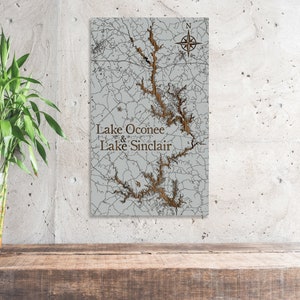 May include: A wooden map of Lake Oconee and Lake Sinclair in Georgia. The map is carved with a light gray background and brown lines representing the lakes and surrounding areas. The text "Lake Oconee & Lake Sinclair" is carved in brown letters at the bottom of the map.