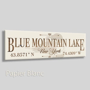 May include: A white canvas sign with brown text that reads "Blue Mountain Lake, New York 43.8571° N 74.4309° W". The sign features a silhouette of a loon.