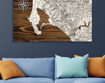 May include: A wooden wall art piece featuring a topographic map of Bodega Bay, California. The map is in black and white and includes a compass rose. The text "Bodega Bay, California" is printed in brown cursive lettering.