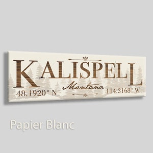 May include: A white canvas sign with brown text that reads "KALISPELL Montana 48.1920° N 114.3168° W". The text is surrounded by a silhouette of pine trees.