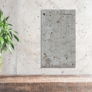 May include: A wood map of Wheaton, Illinois. The map is in a light gray color with brown lines and text. The text "Wheaton, Illinois" is in the top right corner of the map.