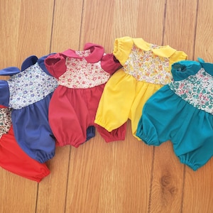 Peut inclure: Cinq barboteuses à fleurs pour poupée de différentes couleurs : rouge, bleu, jaune, vert et turquoise. Chaque barboteuse a un col blanc et des manches courtes.