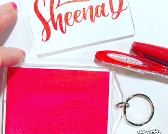 Könnte beinhalten: Ein klarer Acryl-Schlüsselanhänger mit einem hellrosa Einsatz. Eine weiße Karte mit dem Namen "Sheena D" in roter Schrift befindet sich über dem Schlüsselanhänger. Ein roter Stift und die Worte "Hand Lettered & Loved" sind ebenfalls sichtbar.