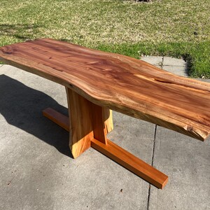 May include: A wooden table with a live edge top and a unique, modern, X-shaped base. The table is made of natural wood with a rich brown color.