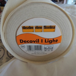May include: A roll of Decovil I Light fusible interfacing, a white fabric used for stabilizing and strengthening fabrics. The label is orange with black text and a barcode. It reads "Decovil I Light", "121", "4008983 212249", "90 cm x 15 m", "42% PES, 35% CV, 23% PA".