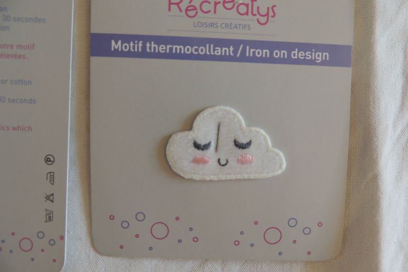 May include: White iron-on patch featuring a smiling cloud design. The cloud has pink cheeks and closed eyes with eyelashes. The text "Motif thermocollant / Iron on design" is printed on the packaging.