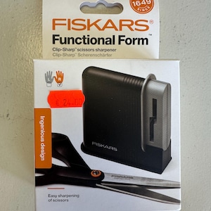 May include: A Fiskars Functional Form scissors sharpener. The black sharpener is on a white box with the Fiskars logo and text. The box also shows a pair of scissors and the text "Easy sharpening of scissors."