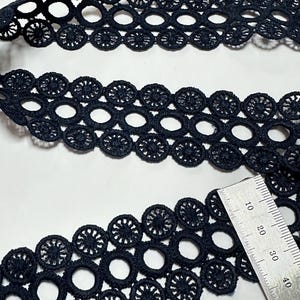 May include: Close-up of black lace trim with a repeating circular pattern. The trim is made of a soft, woven material and is ideal for sewing and crafting projects. A ruler is included in the image to show the size of the lace.