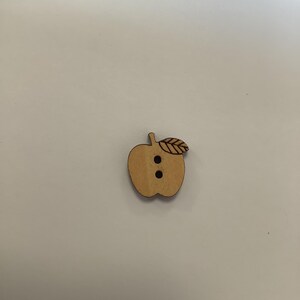 Può includere: Un bottone di legno a forma di mela con un dettaglio a foglia. Il bottone è marrone chiaro con due piccoli fori rotondi per cucire. Il bottone a forma di mela è un materiale artigianale per cucire, lavorare a maglia o altri progetti fai-da-te.