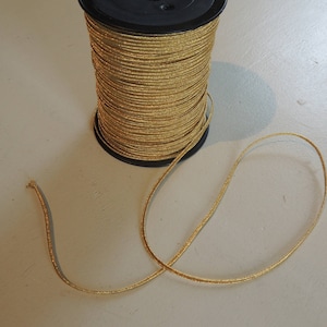 Può includere: Un rocchetto di cordoncino metallico dorato con un rocchetto di plastica nero. Il cordoncino è srotolato e disposto in un anello.