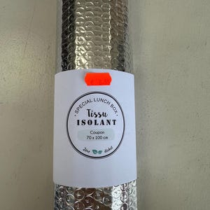 Puede incluir: Un tejido aislante plateado enrollado, etiquetado como "Special Lunch Box. Tissu Isolant". La etiqueta indica un tamaño de cupón de 70 x 100 cm. El tejido está diseñado para mantener los alimentos a una temperatura estable.