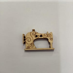 Könnte beinhalten: Ein kleiner, hölzerner Nähmaschinen-Ausschnitt mit dekorativen Wirbeln. Das lasergeschnittene Design zeigt eine detaillierte Kontur einer Vintage-Nähmaschine, perfekt für Bastelprojekte. Das Holz hat eine natürliche, hellbraune Farbe.