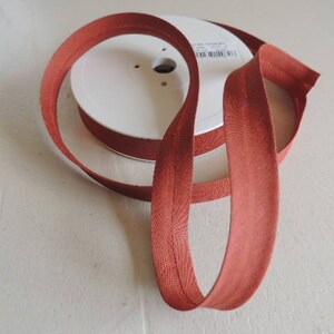 Pode incluir: Uma bobina de fita de vi&eacute;s cor de ferrugem, um tipo de acabamento de tecido usado para finalizar as bordas de roupas e projetos de decora&ccedil;&atilde;o de casa.