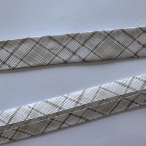 May include: Close-up of two pieces of fabric trim. The trim has a white background with a brown plaid pattern. The trim is folded over, creating a layered effect. The fabric appears to be woven.