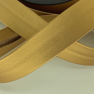 Puede incluir: Cinta de cuero dorada con superficie texturizada. La cinta está enrollada y anudada.