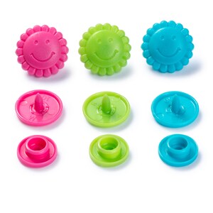 Puede incluir: Conjunto de nueve botones de plástico con un diseño de cara sonriente. Los botones son de tres colores: rosa, verde y azul. Cada color tiene tres botones: un botón grande con una cara sonriente, un botón más pequeño con un centro elevado y un botón plano.