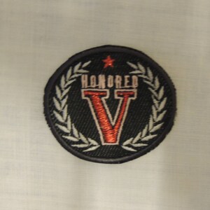 Peut inclure: Patch brodé noir et blanc avec un "V" rouge et le mot "HONORED" en lettres rouges. Le patch est entouré d'une couronne blanche.