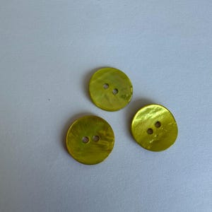Puede incluir: Tres botones redondos amarillos con dos agujeros cada uno. Los botones tienen un brillo nacarado y están dispuestos sobre una superficie gris claro. Estos botones son adecuados para proyectos de costura y manualidades.