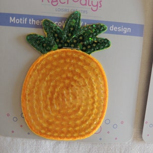 Può includere: Un toppa termoadesiva a forma di ananas gialla con paillettes verdi per le foglie. La toppa è su uno sfondo bianco con il testo "Motif there coll/ design" sopra.