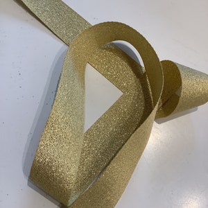 May include: Gold ribbon with a herringbone pattern and a subtle glitter finish. The ribbon is folded and twisted, showing the texture and shimmer.