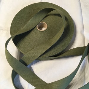 May include: A roll of olive green webbing, a type of heavy-duty fabric tape, is shown on a white surface. The webbing is rolled up and tied in a knot.