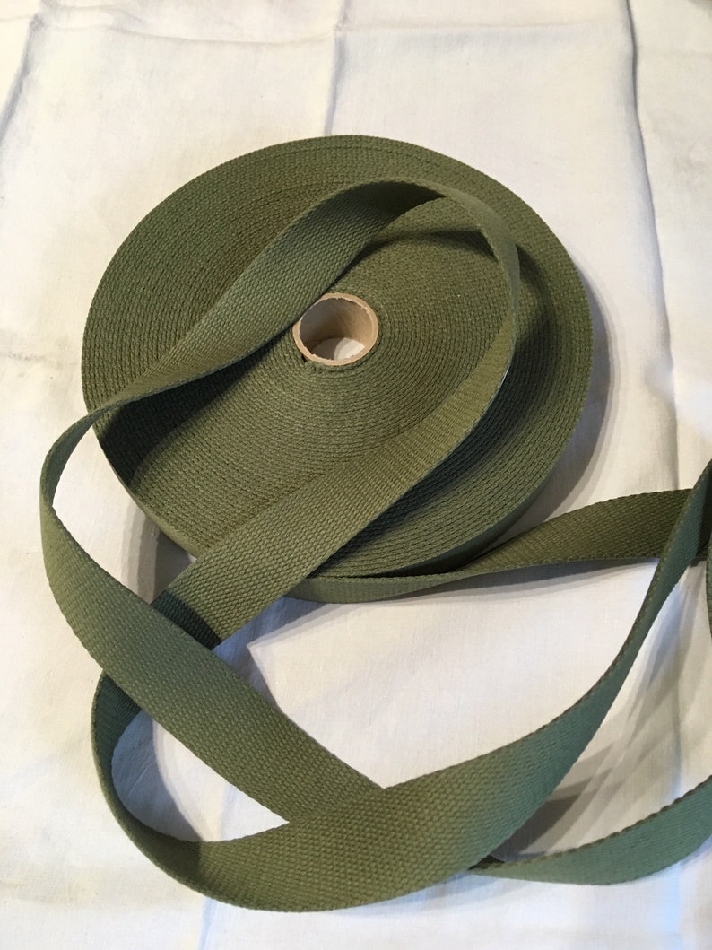 May include: A roll of olive green webbing, a type of heavy-duty fabric tape, is shown on a white surface. The webbing is rolled up and tied in a knot.
