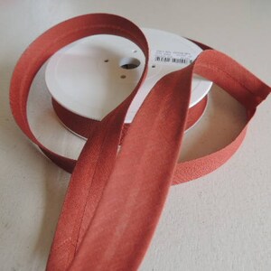 Pode incluir: Uma bobina de guarni&ccedil;&atilde;o de tecido cor de ferrugem com um r&oacute;tulo branco. A guarni&ccedil;&atilde;o &eacute; dobrada e enrolada na bobina.