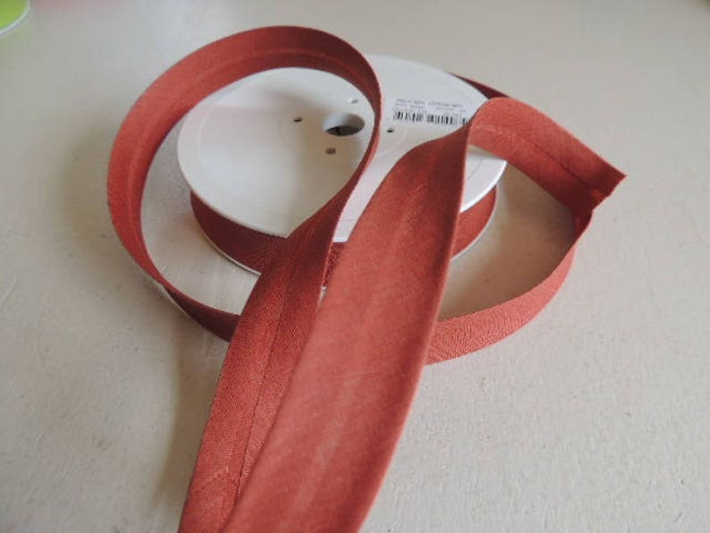 Pode incluir: Uma bobina de guarni&ccedil;&atilde;o de tecido cor de ferrugem com um r&oacute;tulo branco. A guarni&ccedil;&atilde;o &eacute; dobrada e enrolada na bobina.