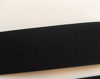 May include: Close-up of a strip of black hook and loop fastener material. The material has a textured surface with small, closely spaced loops. The background is plain white.