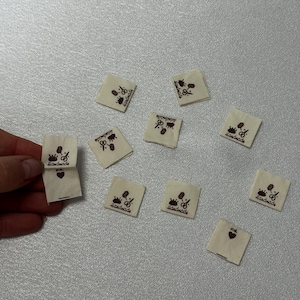 Peut inclure: Plusieurs petites étiquettes carrées en tissu blanc cassé avec des textes et des graphiques bruns. Chaque étiquette porte le mot "Handmade" avec un motif de ciseaux et de couronne. Certaines étiquettes comportent également un symbole de cœur.
