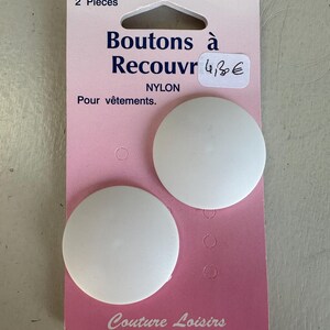 May include: Two white nylon buttons, each 1.5 inches in diameter, are displayed on a pink and white card. The card has text in French, including "Boutons à Recouvr" and "Pour vêtements."