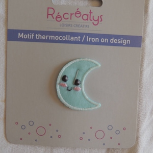 May include: A light blue iron-on patch with a crescent moon design. The moon has a smiling face with black eyes and pink cheeks. The text "Motif thermocollant / Iron on design" is printed on the packaging.