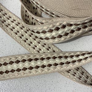 Peut inclure: Gros plan sur un rouleau de ruban beige tissé avec un motif répétitif de losanges marron et blanc cassé. Le ruban est tissé serré et semble être fait d'un matériau durable, adapté à divers projets d'artisanat.