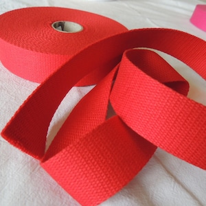 Può includere: Immagine ravvicinata di un nastro di cotone rosso, un tipo di tessuto piatto tessuto. Il nastro è arrotolato e piegato in modo da mostrare la trama del tessuto.