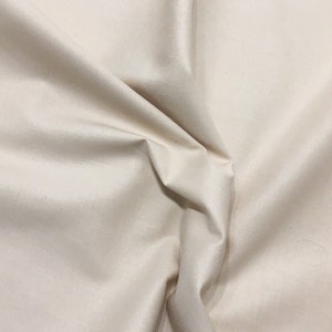 Könnte beinhalten: Nahaufnahme eines beigefarbenen Stoffes mit weichen Falten und dezentem Glanz. Das Material wirkt glatt und möglicherweise seidig, wobei Lichtreflexe und Schatten Tiefe verleihen. Der Stoff ist einfarbig und eignet sich für verschiedene Näh- und Bastelprojekte.